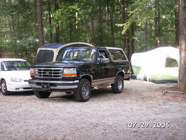Black Ford Bronco SUV with tan lower trim parked at a wooded gravel campsite next to a white car and tents