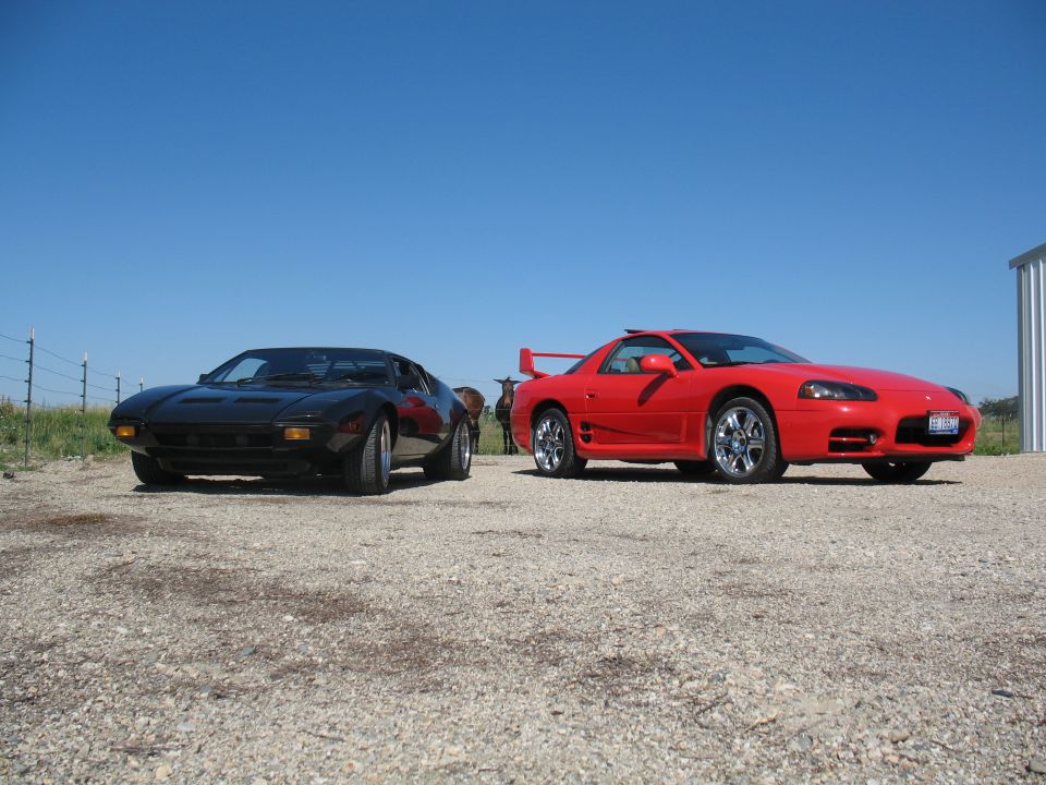 Black De Tomaso Pantera next to a red sports coupe on gravel under blue sky