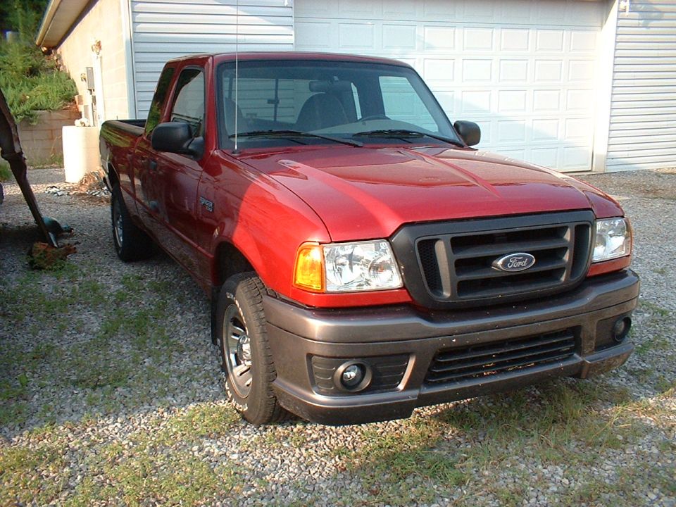Red Ford Ranger extended cab pickup on gravel by garage, front three-quarter view