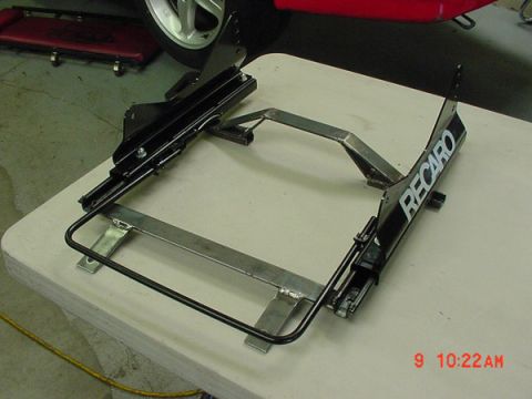 1993 Ford Mustang - A close-up view of a black Recaro seat bracket assembly, with a portion of a red vehicle and wheel in the background.