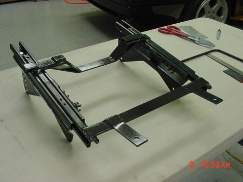 1993 Ford Mustang - A close-up view of a black metal seat mounting bracket assembly, possibly for a car, rests on a workbench.