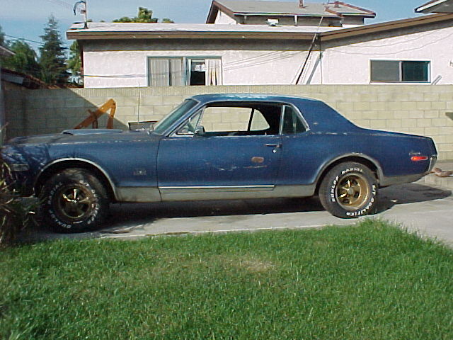 Side view of a worn dark blue 1968 Mercury Cougar coupe on gold wheels in a driveway