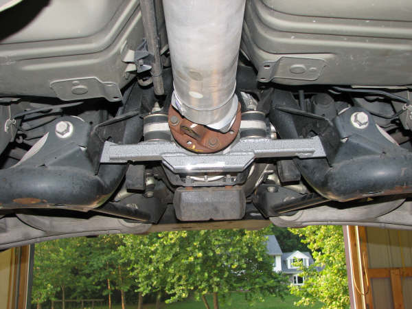 Underside view of a vehicle's rear axle, driveshaft, and exhaust pipe. Supermotors.net logo visible