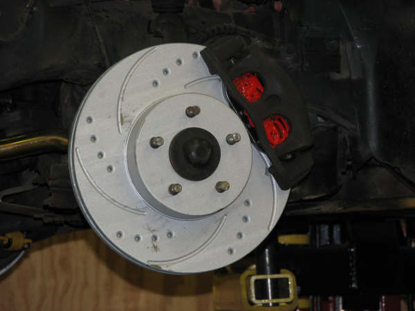 Close-up of a car's front brake rotor and caliper assembly, showing drilled rotor and red brake pads