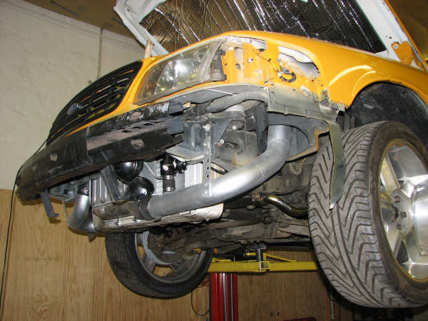 Yellow car front end with hood open, showing custom exhaust pipes and intercooler