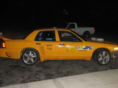 2001 Mercury Grand Marquis - Modified yellow taxi with racing decals and a hood scoop is parked next to a white pickup truck.