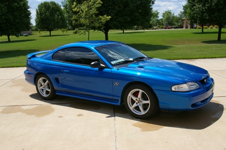 1998 Ford Mustang - A blue 1998 Ford Mustang is parked on a concrete driveway with a grassy lawn and trees in the background.