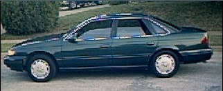1994 Ford Taurus - A dark green sedan is parked on the side of the road with a tree and bushes in the background.