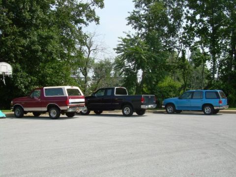 1992 Ford Bronco - Three vehicles are parked in a lot with trees and a basketball hoop in the background.