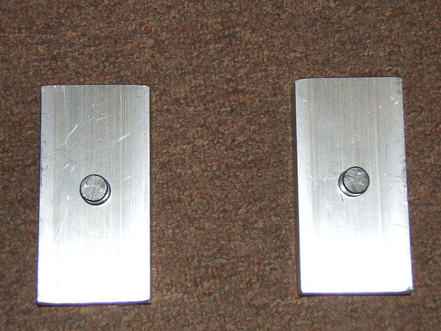two rectangular metal plates, each with a single bolt in the center, lying on a brown carpet