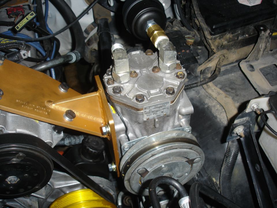 Engine bay shot of an AC compressor with a gold bracket and hoses attached