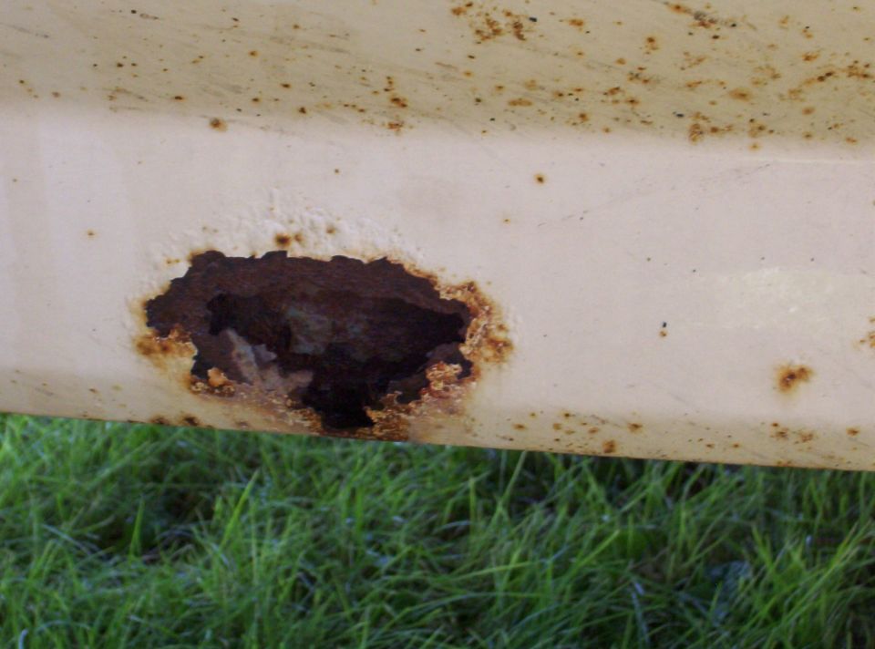 Close-up of a rust hole with brown corrosion on a tan painted body panel, grass visible below