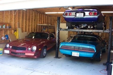 1970 Chevrolet Camaro - Three classic muscle cars are parked in a garage, with one car elevated on a lift.