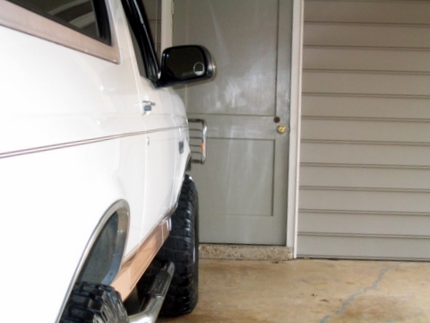 1996 Ford Bronco - The side of a white SUV is visible, parked in front of a closed door and garage.