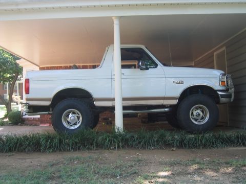1996 Ford Bronco - Passenger-side profile of a white full-size Ford Bronco with a tan lower two-tone stripe and the hardtop removed, parked under a carport. I…