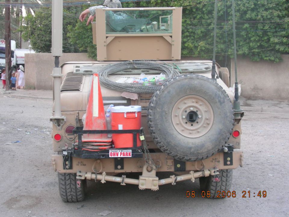 Rear view of a tan military truck with a spare tire, orange traffic cone, and cooler mounted on the back. Text on a sticker reads 'OHV PARK'