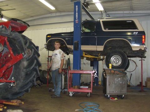 1986 Ford Bronco - A mechanic stands next to a blue and tan Ford Bronco being serviced on a lift in a garage.