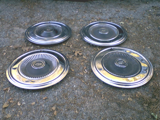 Four chrome hubcaps with ribbed centers arranged on gravel ground