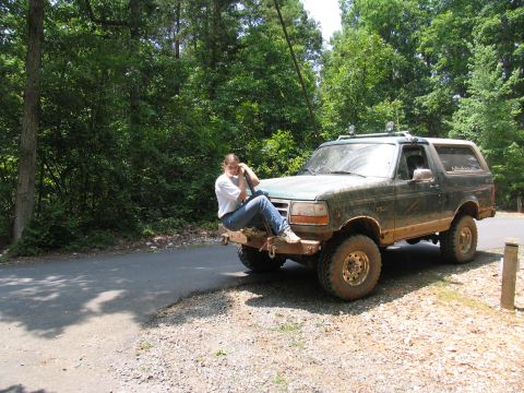 1996 Ford Bronco - A person sits on the front bumper of a muddy Ford Bronco parked on the side of a wooded road.