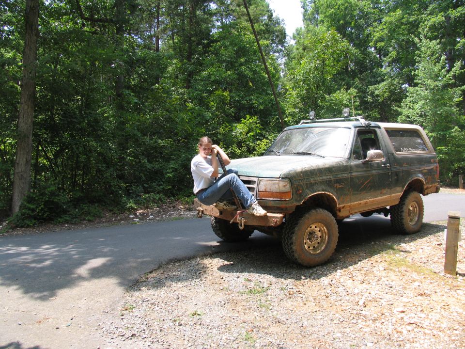 Person sitting on front bumper of a dark green Ford Bronco parked on a dirt road