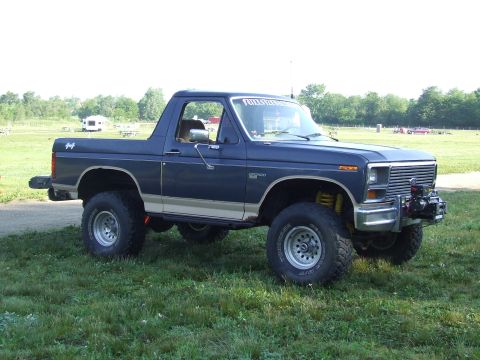 1986 Ford Bronco - This lifted Ford Bronco sits on a grassy field with trees in the background and a camper visible in the distance.