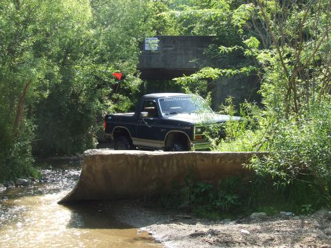 1986 Ford Bronco - A Ford Bronco navigates a rocky creek bed under a bridge, surrounded by dense greenery.