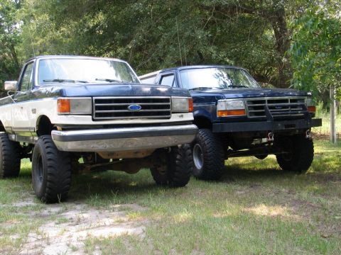 1989 Ford F-150 - Two lifted Ford trucks, one appearing to be a 1989 F-150, are parked side-by-side outdoors.