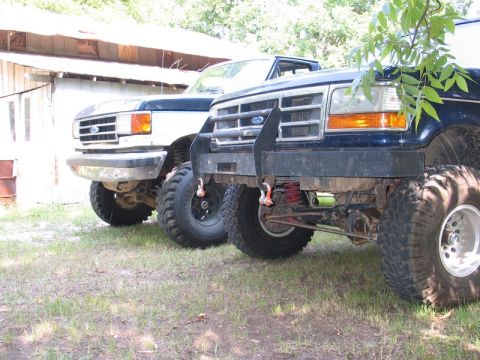 1989 Ford F-150 - Two lifted Ford trucks are parked side-by-side in front of a rustic building.