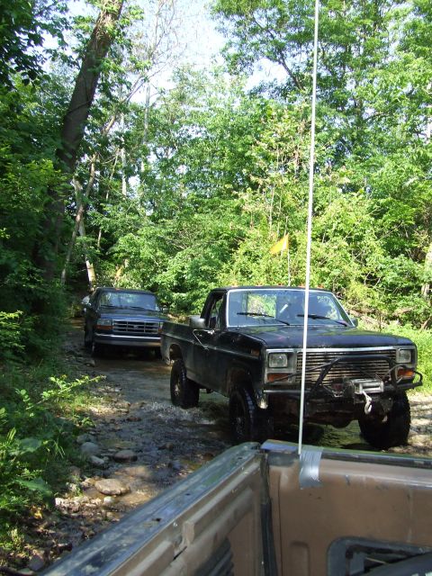 1986 Ford Bronco - Two trucks navigate a muddy path through a dense forest on a sunny day.