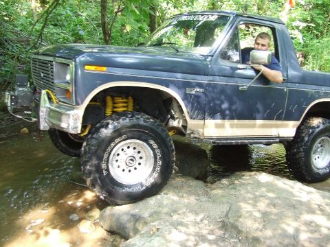 1986 Ford Bronco - A man drives a modified Ford Bronco through a rocky stream in a wooded area.