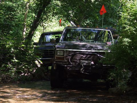 1986 Ford Bronco - Two off-road SUVs navigate a wooded trail with orange flags marking their presence.