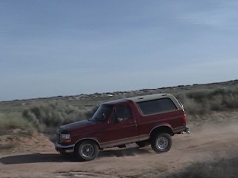 1994 Ford Bronco - Red 1994 Ford Bronco kicks up dust on a dirt road under a clear blue sky.