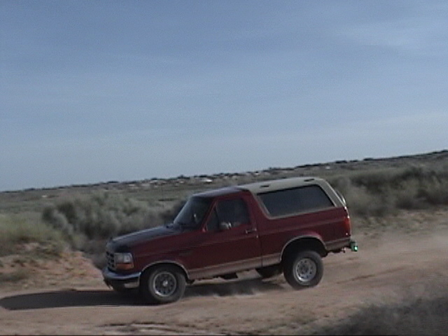 1994 Ford Bronco driving on a dirt road with brush and hills in background