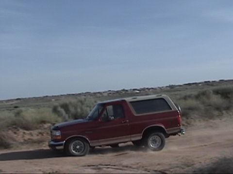 1994 Ford Bronco - A red Ford Bronco with a tan top is seen driving on a dirt road under a clear blue sky.