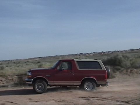 1994 Ford Bronco - A red 1994 Ford Bronco kicks up dust as it drives on a dirt road under a clear blue sky.
