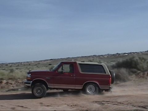 1994 Ford Bronco - This red Ford Bronco is seen from the side, driving on a dusty road with a spare tire on the back.