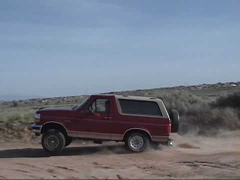 1994 Ford Bronco - A red Ford Bronco drives off-road, kicking up dust on a sunny day.