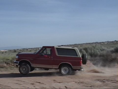 1994 Ford Bronco - A red Ford Bronco drives off-road, its tires kicking up dust under a clear blue sky.