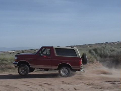 1994 Ford Bronco - A red 1994 Ford Bronco drives off-road, kicking up a cloud of dust.