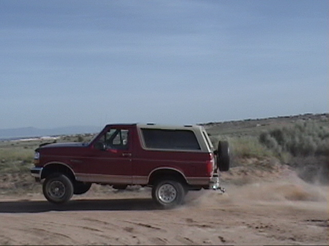Red 1994 Ford Bronco driving on a dirt road, kicking up dust. Spare tire mounted on the rear