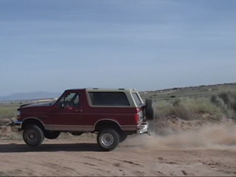 1994 Ford Bronco - A red Ford Bronco drives on a dirt road, creating a dust cloud behind it under a clear blue sky.