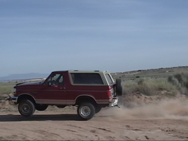 Red Ford Bronco with a tan top, kicking up dust on a dirt road. Spare tire mounted on the rear