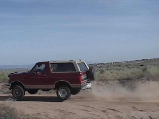 Red 1994 Ford Bronco driving on a dirt road, kicking up dust. Spare tire mounted on rear