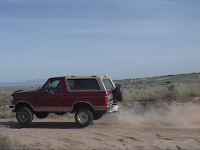 Red 1994 Ford Bronco driving on a dirt road, kicking up dust. Spare tire mounted on the back