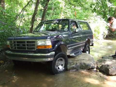 1986 Ford Bronco - Ford Bronco driving off-road through a creek bed with trees in the background.
