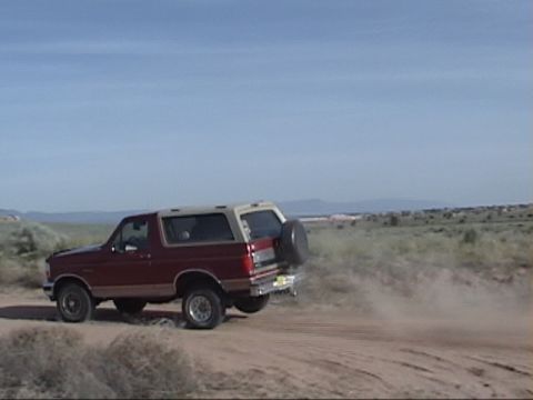 1994 Ford Bronco - A red and tan SUV drives off-road, kicking up dust under a clear blue sky.