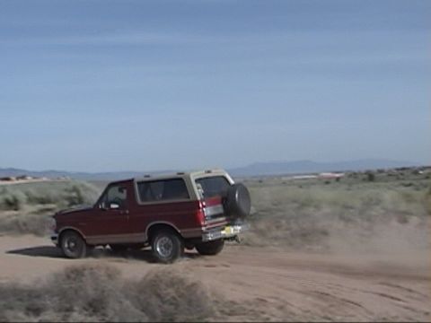 1994 Ford Bronco - Red and tan 1994 Ford Bronco kicks up dust on a dirt road under a clear blue sky.
