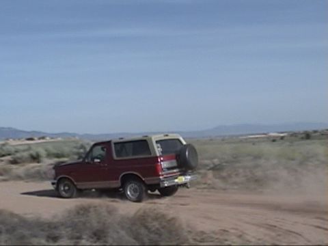 1994 Ford Bronco - A red SUV kicks up dust as it drives along a dirt road under a clear blue sky.