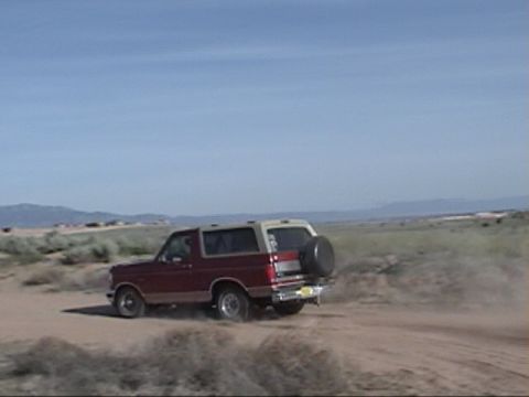 1994 Ford Bronco - A Ford Bronco kicks up dust as it drives across a dry, open landscape under a clear blue sky.