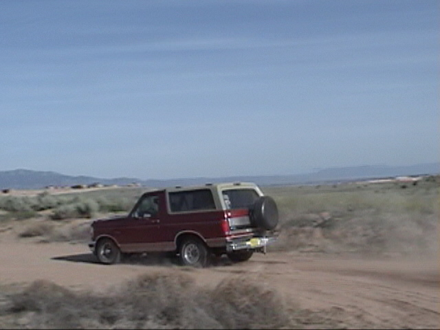 Red and tan Ford Bronco with a spare tire on the back, driving on a dirt road